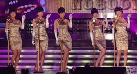 Five women with beehive hairstyles wearing matching golden sheath dresses and elbow-length gloves