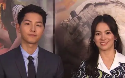 Actors Song Joong-ki and Song Hye-kyo pose side-by-side in front of a promotional backdrop for "Descendants of the Sun"
