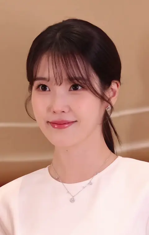 Foto jarak dekat penyanyi dan aktris Korea Selatan, IU, yang tersenyum lembut dengan poni samping dan rambut diikat ke belakang. Dia mengenakan atasan putih, anting-anting perak kecil, dan kalung perak tipis dengan liontin kecil. Latar belakangnya adalah interior berwarna krem yang buram.