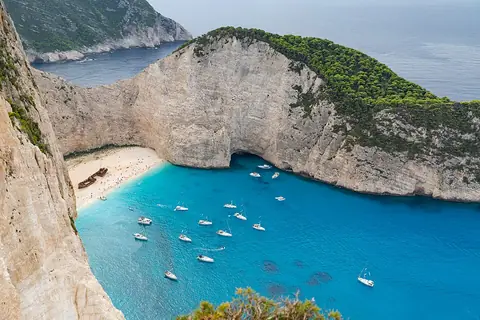 A high-angle view of a turquoise bay in Zakynthos with white sand, a shipwreck, and numerous sailboats anchored between steep limestone cliffs.