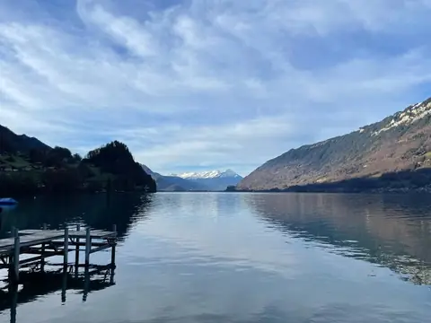 Panoramic view of a tranquil Swiss lake surrounded by mountains, with a wooden dock in the foreground and snow-capped peaks under a partly cloudy sky in the distance.