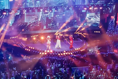 An image of the stage taken at a distance so part of the floor crowd is visible. The singers are wearing white and red on a stage with a Christmas theme.
