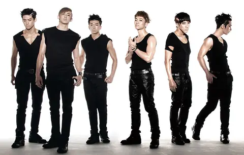 Six men wearing form-fitting black sleeveless shirts, leather pants, and combat boots. They have prominent eye makeup and each has a different gelled hairstyle.